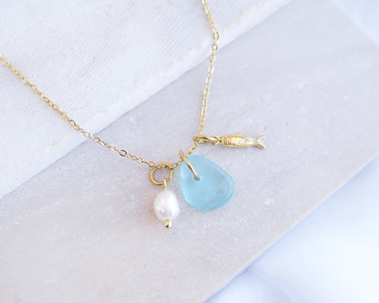 Close up of a necklace with a tiny sardine fish pendant, turquoise blue sea glass and freshwater pearl pendant on a white stone background. Made with gold stainless steel chain.