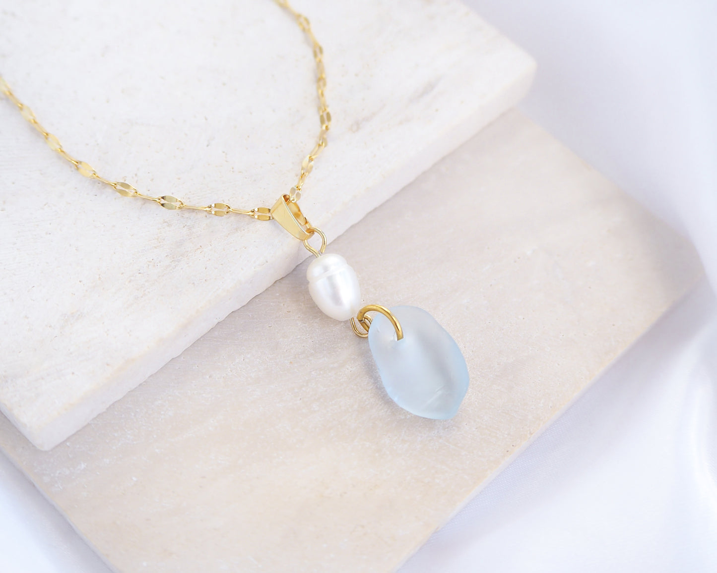 Necklace with a pearl and teardrop blue seaglass pendant on stone displayed., Coastal jewelry with freshwater pearl necklace. Made with light blue sea glass and 18k gold stainless steel. Ocean-inspired jewelry, beach necklace, ocean treasure necklace