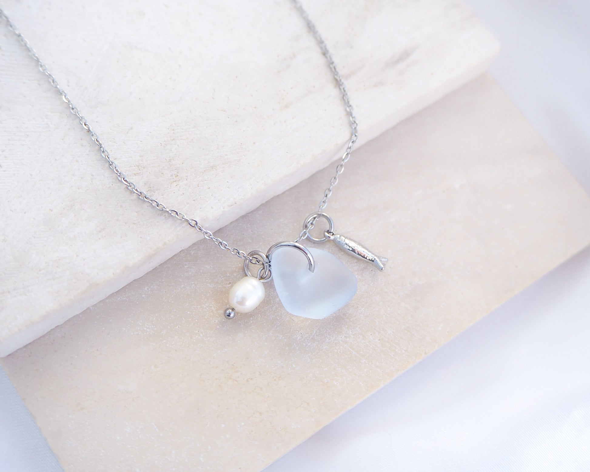Close up of a necklace with a tiny sardine fish pendant, light blue sea glass and freshwater pearl pendant on a white stone background. Made with silver stainless steel chain.