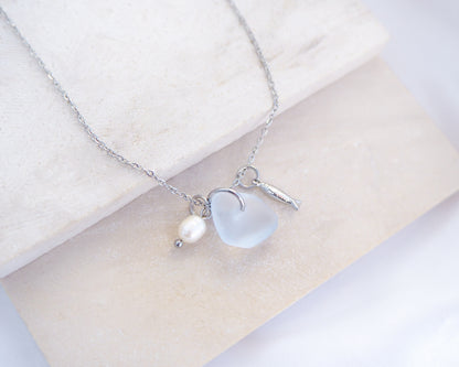 Close up of a necklace with a tiny sardine fish pendant, light blue sea glass and freshwater pearl pendant on a white stone background. Made with silver stainless steel chain.