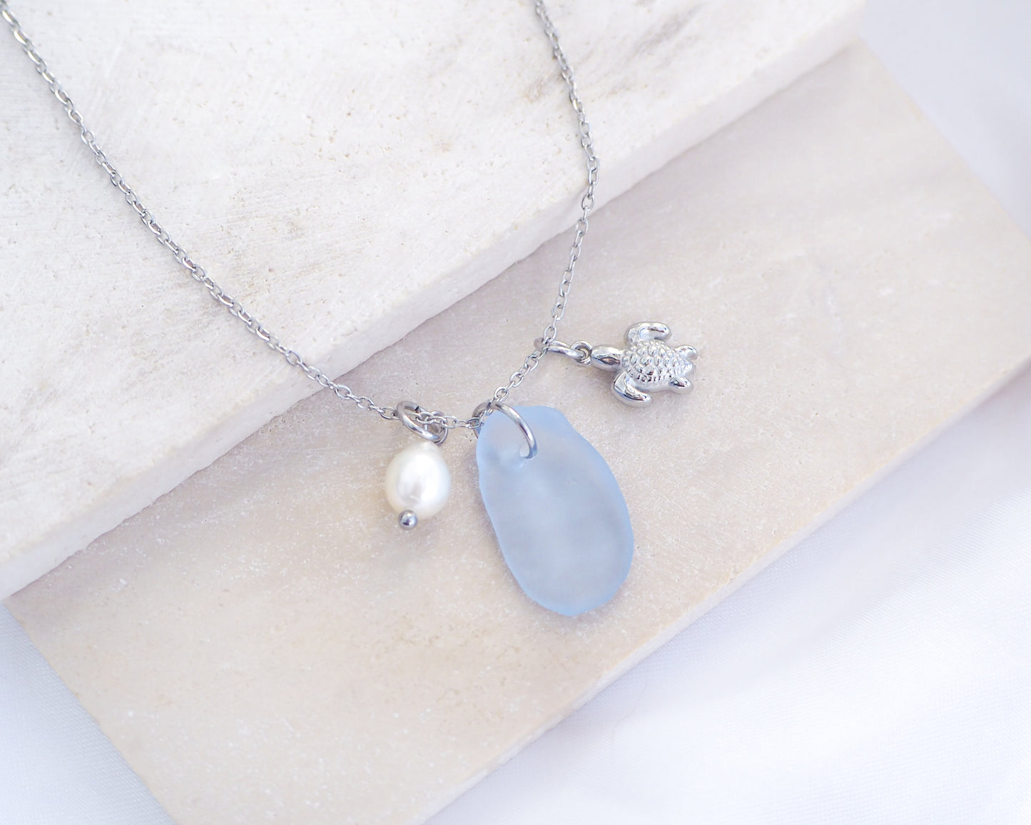 A silver necklace with a tiny turtle pendant, light blue sea glass and freshwater pearl pendant on a white stone background. Made with silver stainless steel chain.  