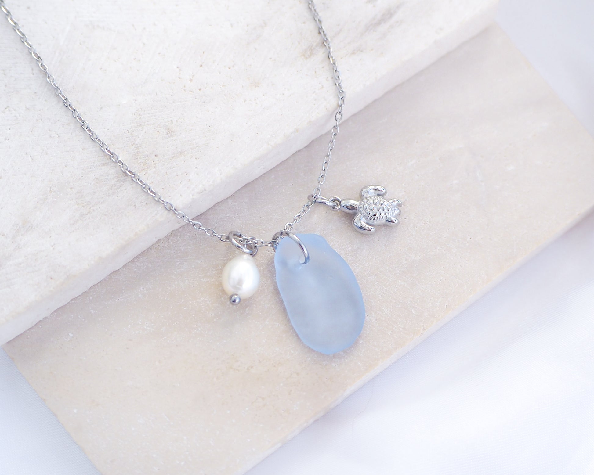 A silver necklace with a tiny turtle pendant, light blue sea glass and freshwater pearl pendant on a white stone background. Made with silver stainless steel chain.  
