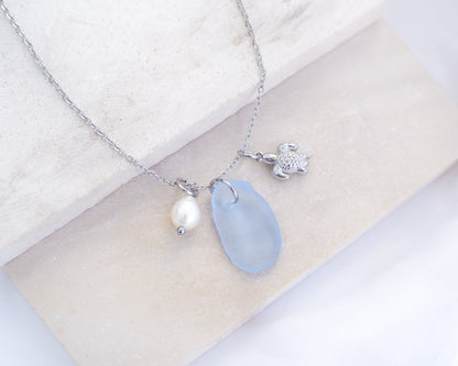 A silver necklace with a tiny turtle pendant, light blue sea glass and freshwater pearl pendant on a white stone background. Made with silver stainless steel chain.  