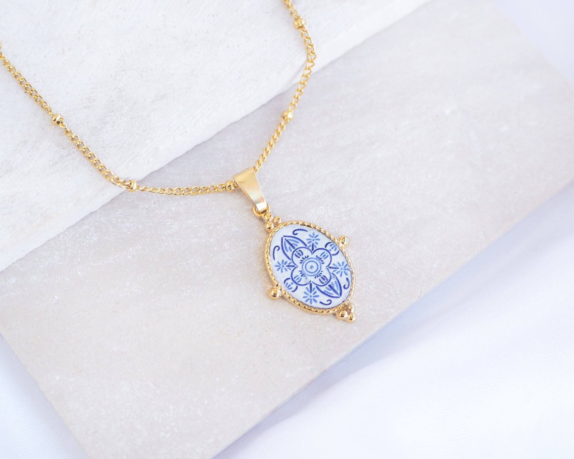 Close up of Blue Portuguese Tile Oval Deluxe Necklaces Blue on stone displayed. Made with 18k gold stainless steel and white blue mosaic pendant inspired by the Portuguese Azulejo design pattern. 