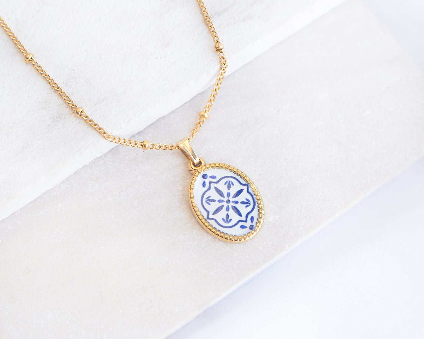 Close up of Blue White Portuguese Tile Oval Necklaces Blue on stone displayed. Made with 18k gold stainless steel and white blue mosaic pendant inspired by the Portuguese Azulejo design pattern. 