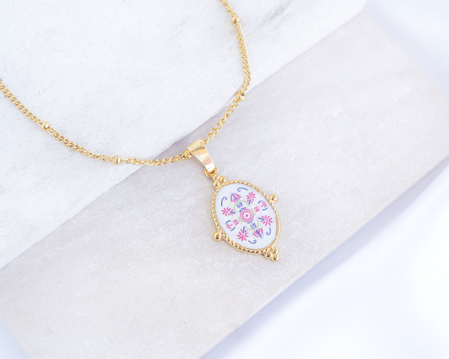 Close up of Pink Blue Portuguese Tile Oval Necklaces Blue on stone displayed. Made with 18k gold stainless steel and white blue mosaic pendant inspired by the Portuguese Azulejo design pattern. 