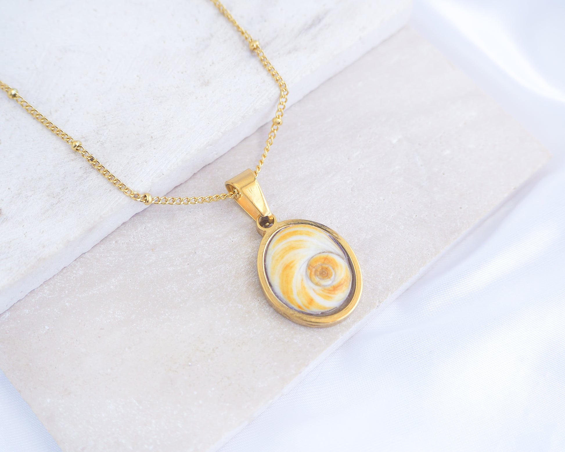 A gold chain necklace with a round pendant featuring a yellow and white spiral pattern, displayed on a light textured surface. Gold Yellow Orange Shiva Eye, Eye of Saint Lucia Gold Necklace on stone displayed, 18k gold stainless steel shiva Yellow Orange eye pendant necklace, Saint Lucy Eye, operculum pendant