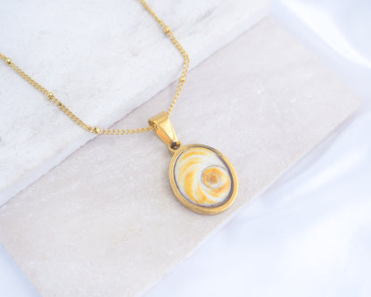 A gold chain necklace with a round pendant featuring a yellow and white spiral pattern, displayed on a light textured surface. Gold Yellow Orange Shiva Eye, Eye of Saint Lucia Gold Necklace on stone displayed, 18k gold stainless steel shiva Yellow Orange eye pendant necklace, Saint Lucy Eye, operculum pendant