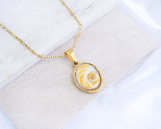 A gold chain necklace with a round pendant featuring a yellow and white spiral pattern, displayed on a light textured surface. Gold Yellow Orange Shiva Eye, Eye of Saint Lucia Gold Necklace on stone displayed, 18k gold stainless steel shiva Yellow Orange eye pendant necklace, Saint Lucy Eye, operculum pendant