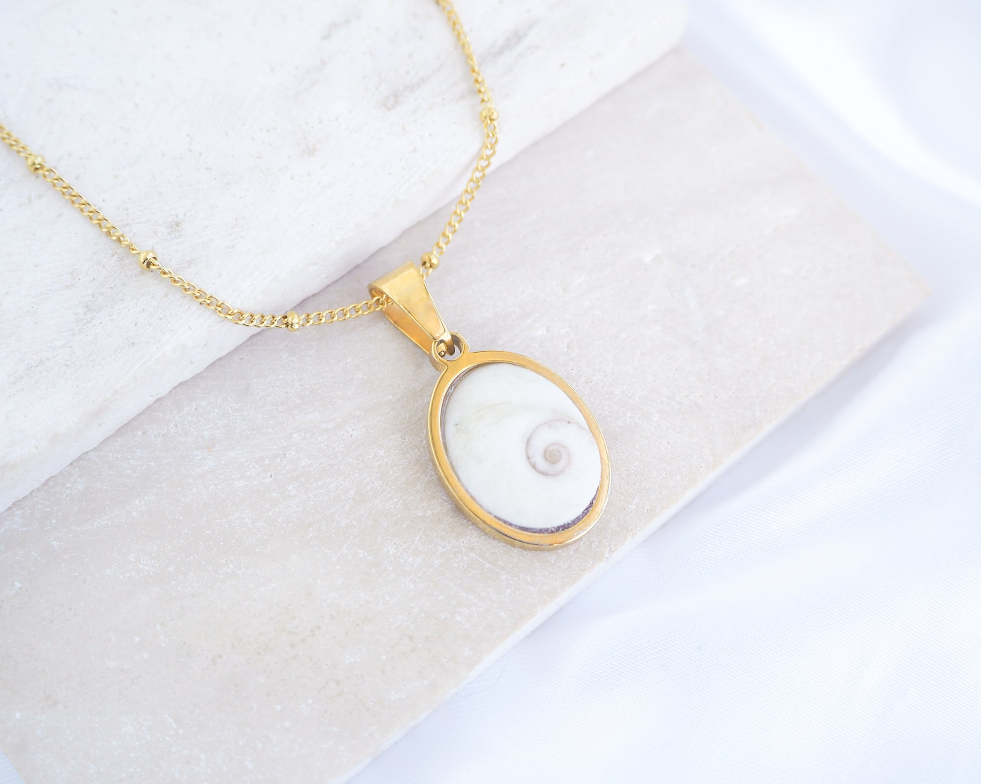 White Eye of Saint Lucia Gold Necklace III on stone displayed, 18k gold stainless steel shiva eye pendant dot chain necklace, Saint Lucy Eye, operculum pendant