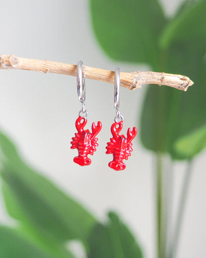 Front view of Red Lobster Silver Earrings with green plant background. Made with silver stainless steel. 