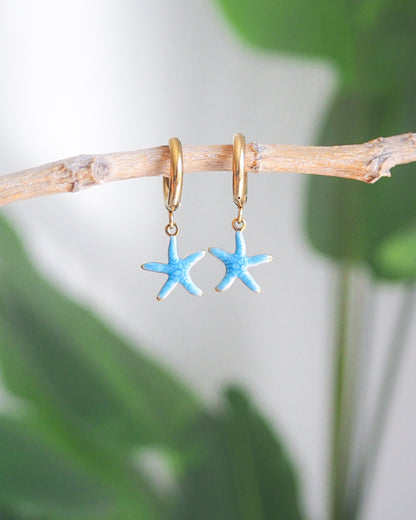 Front view of tiny starfish hoop earrings with green plant background. The starfish pendants are blue. Made with 18k gold stainless steel earring hoops. Beach jewelry for summer days. Minimalistic coastal earrings. Perfect to mix and match with other earrings.