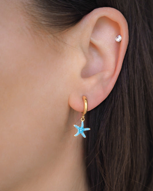 Model wearing a tiny starfish hoop earring in ear. The starfish pendants are blue. Made with 18k gold stainless steel earring hoops. Beach jewelry for summer days. Minimalistic coastal earrings. Perfect to mix and match with other earrings.