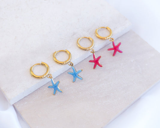 Two pairs of tiny starfish hoop earrings displayed on light stone. The starfish pendants are red and blue. Made with 18k gold stainless steel earring hoops. Beach jewelry for summer days. Minimalistic coastal earrings. Perfect to mix and match with other earrings.