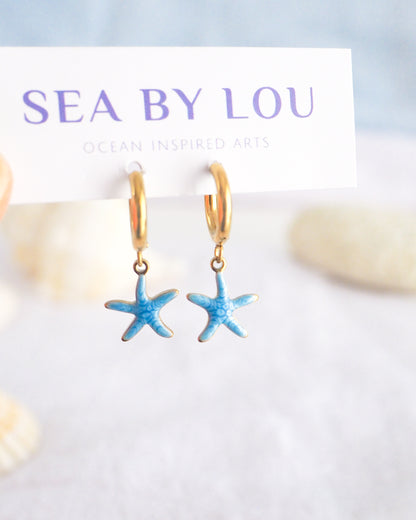 Front view of tiny starfish hoop earrings on white Sea by Lou jewelry card. The starfish pendants are blue. Made with 18k gold stainless steel earring hoops. Beach jewelry for summer days. Minimalistic coastal earrings. Perfect to mix and match with other earrings.