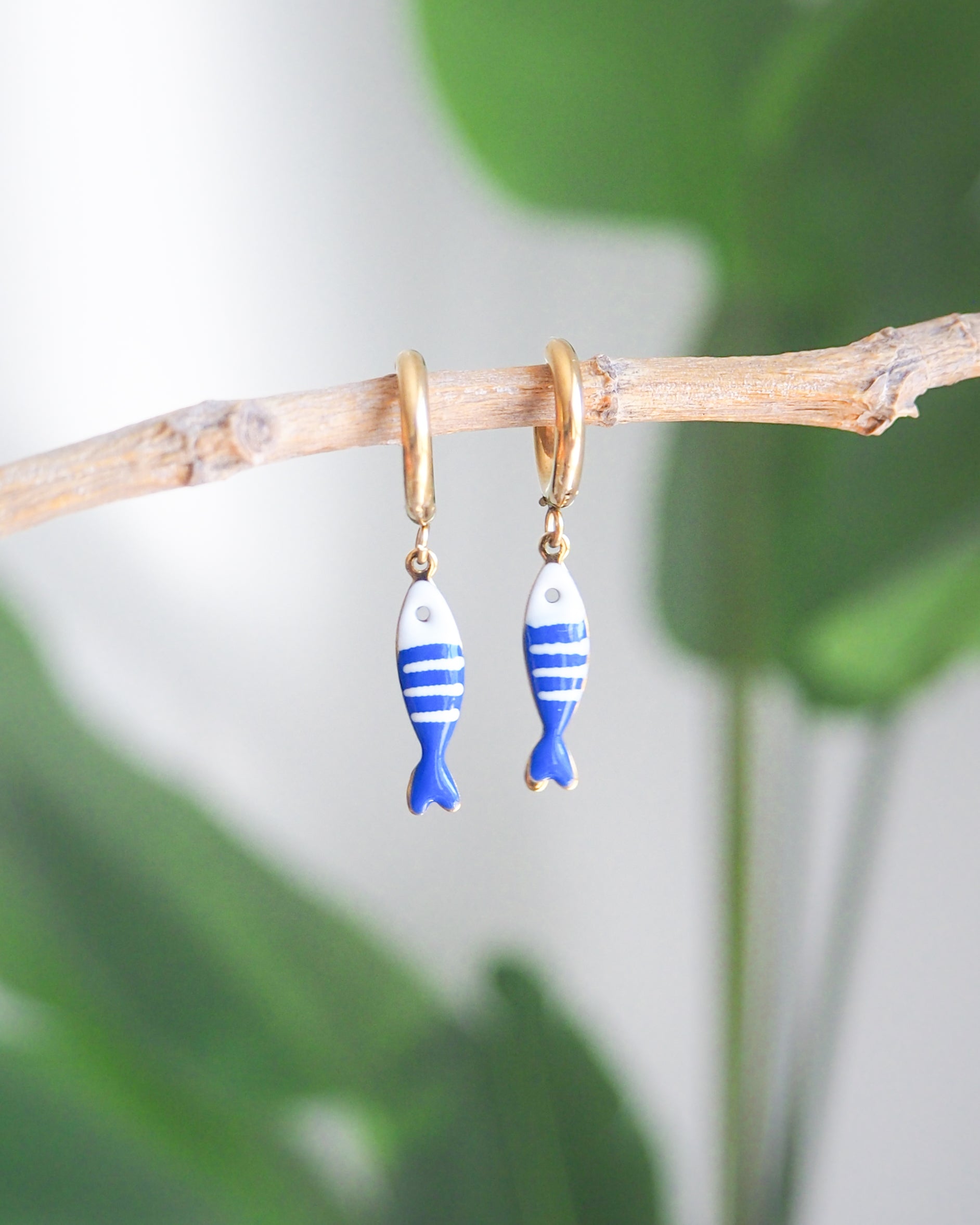 Front view of tiny sardine fish hoop earrings with green plant background. The fish pendant is blue and white striped. Made with 18k gold stainless steel earring hoops. Beautiful gift for Portugal lovers or holiday memory, coastal jewelry from Portugal. Nautical minimalistic jewelry 