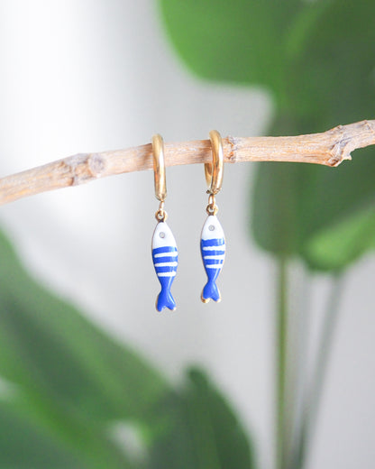 Front view of tiny sardine fish hoop earrings with green plant background. The fish pendant is blue and white striped. Made with 18k gold stainless steel earring hoops. Beautiful gift for Portugal lovers or holiday memory, coastal jewelry from Portugal. Nautical minimalistic jewelry 