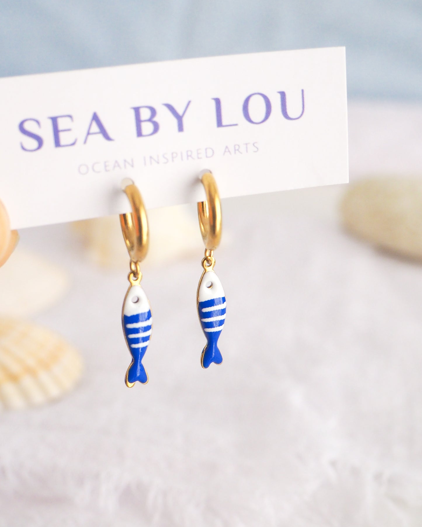 Front view of tiny sardine fish hoop earrings displayed on white Sea by Lou jewelry card. The fish pendant is blue and white striped. Made with 18k gold stainless steel earring hoops. Beautiful gift for Portugal lovers or holiday memory, coastal jewelry from Portugal. Nautical minimalistic jewelry 