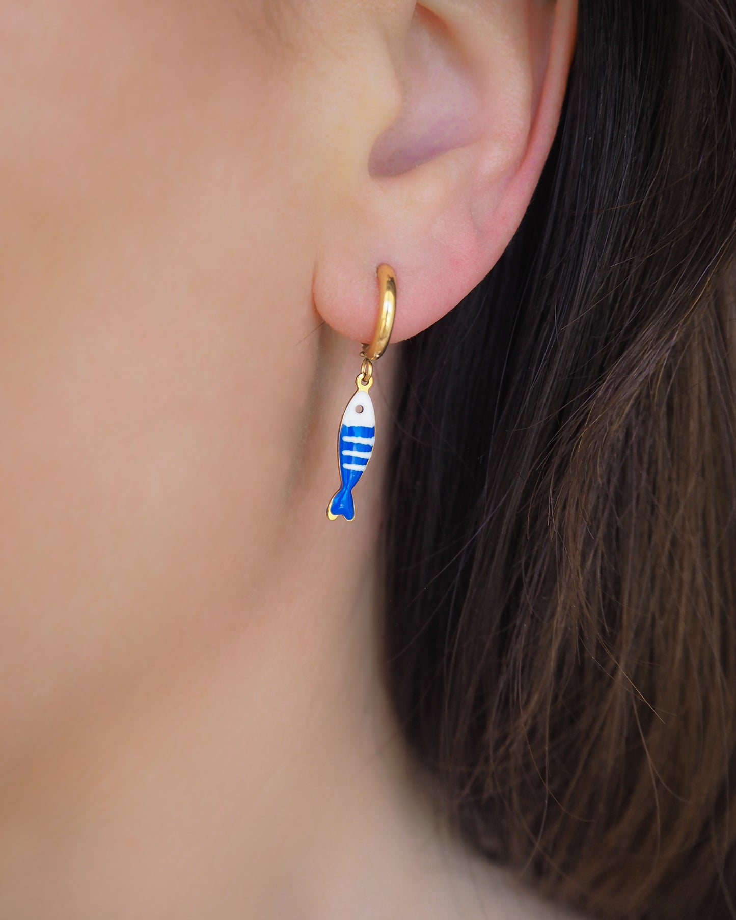 Model wearing a earring with tiny blue white striped sardine fish pendant. Made with 18k gold stainless steel earring hoops. Beautiful gift for Portugal lovers or holiday memory, coastal jewelry from Portugal. Nautical minimalistic jewelry 