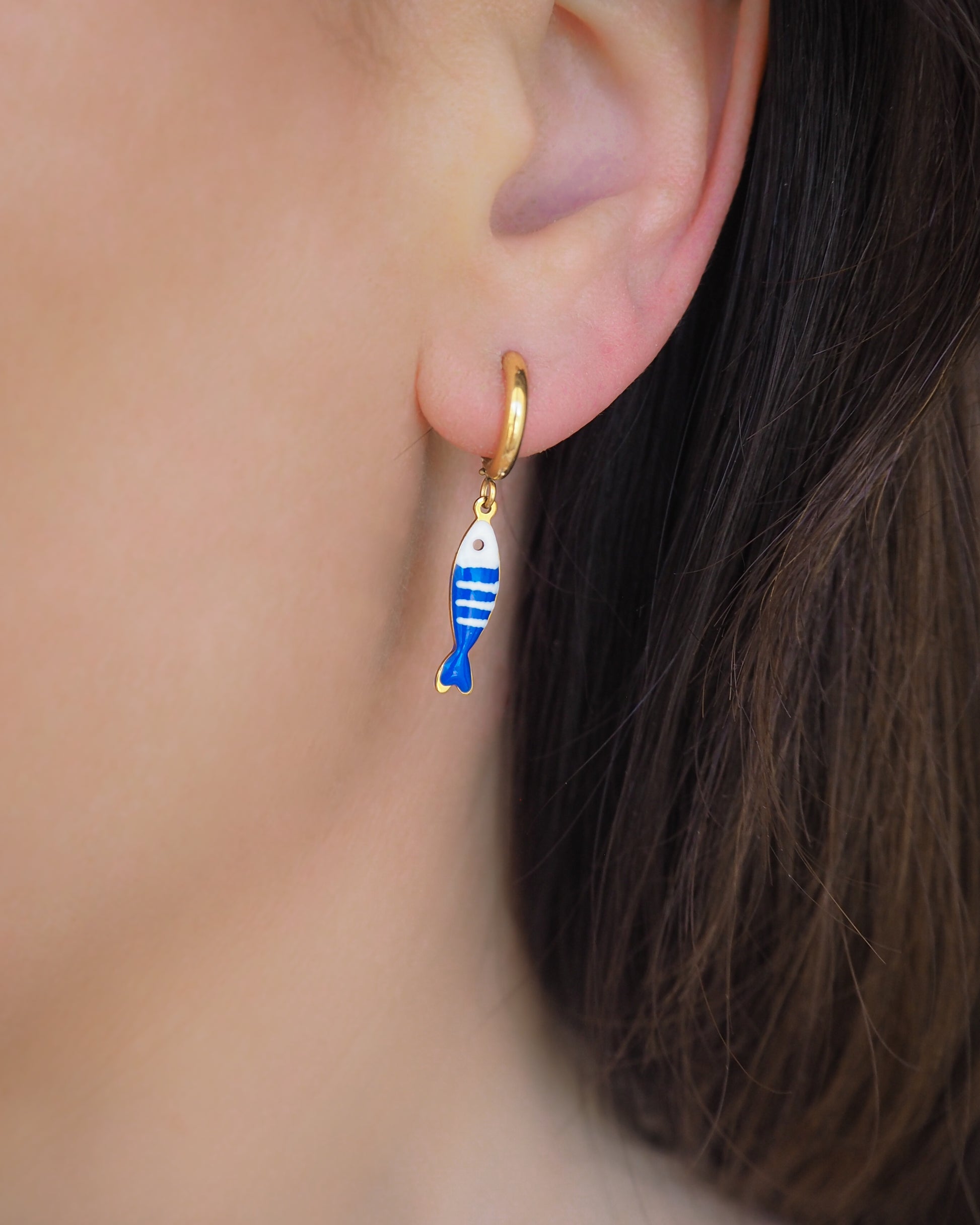 Model wearing a earring with tiny blue white striped sardine fish pendant. Made with 18k gold stainless steel earring hoops. Beautiful gift for Portugal lovers or holiday memory, coastal jewelry from Portugal. Nautical minimalistic jewelry 