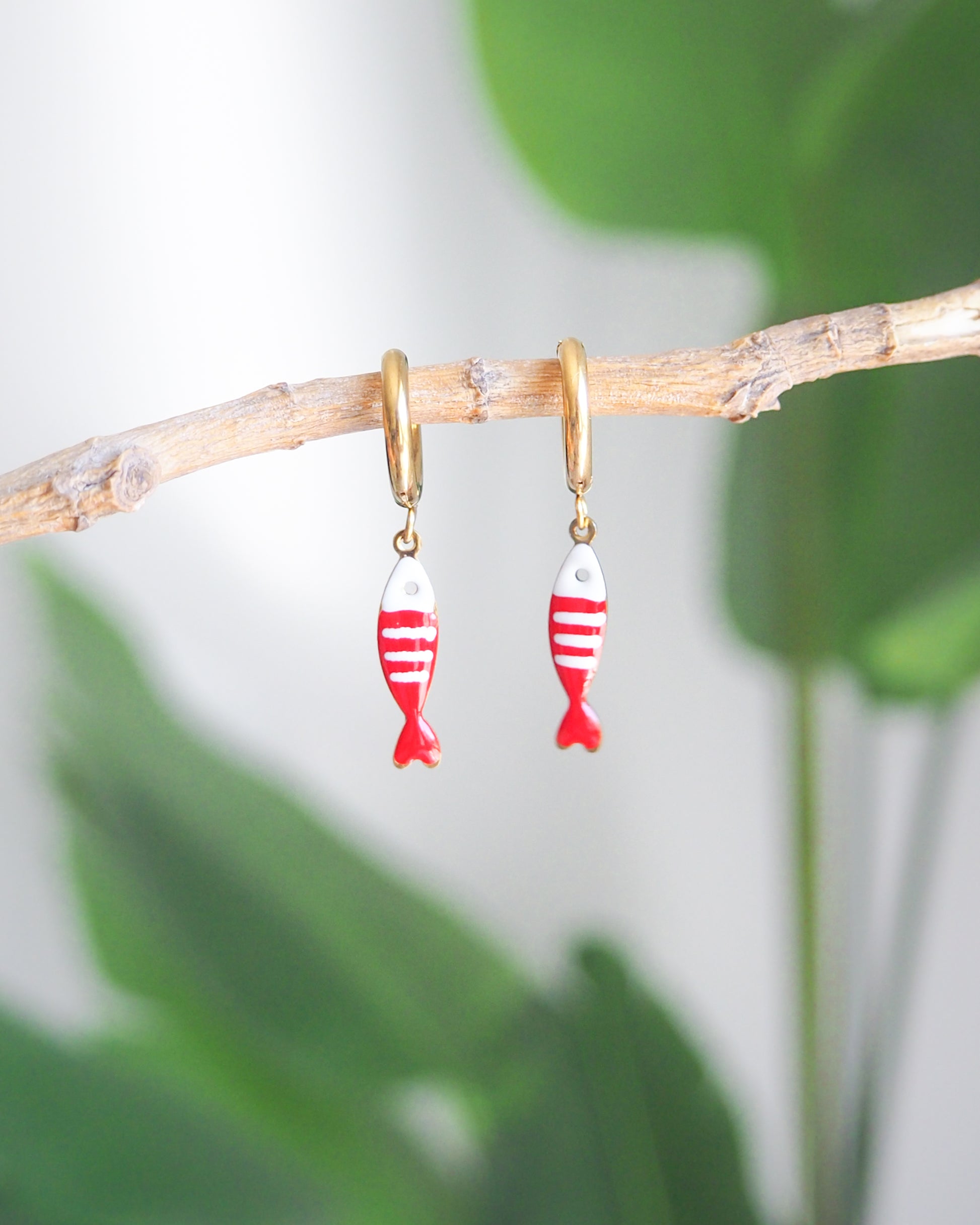 Front view of tiny sardine fish hoop earrings with green plant background. The fish pendant is red and white striped. Made with 18k gold stainless steel earring hoops. Beautiful gift for Portugal lovers or holiday memory, coastal jewelry from Portugal. Nautical minimalistic jewelry 