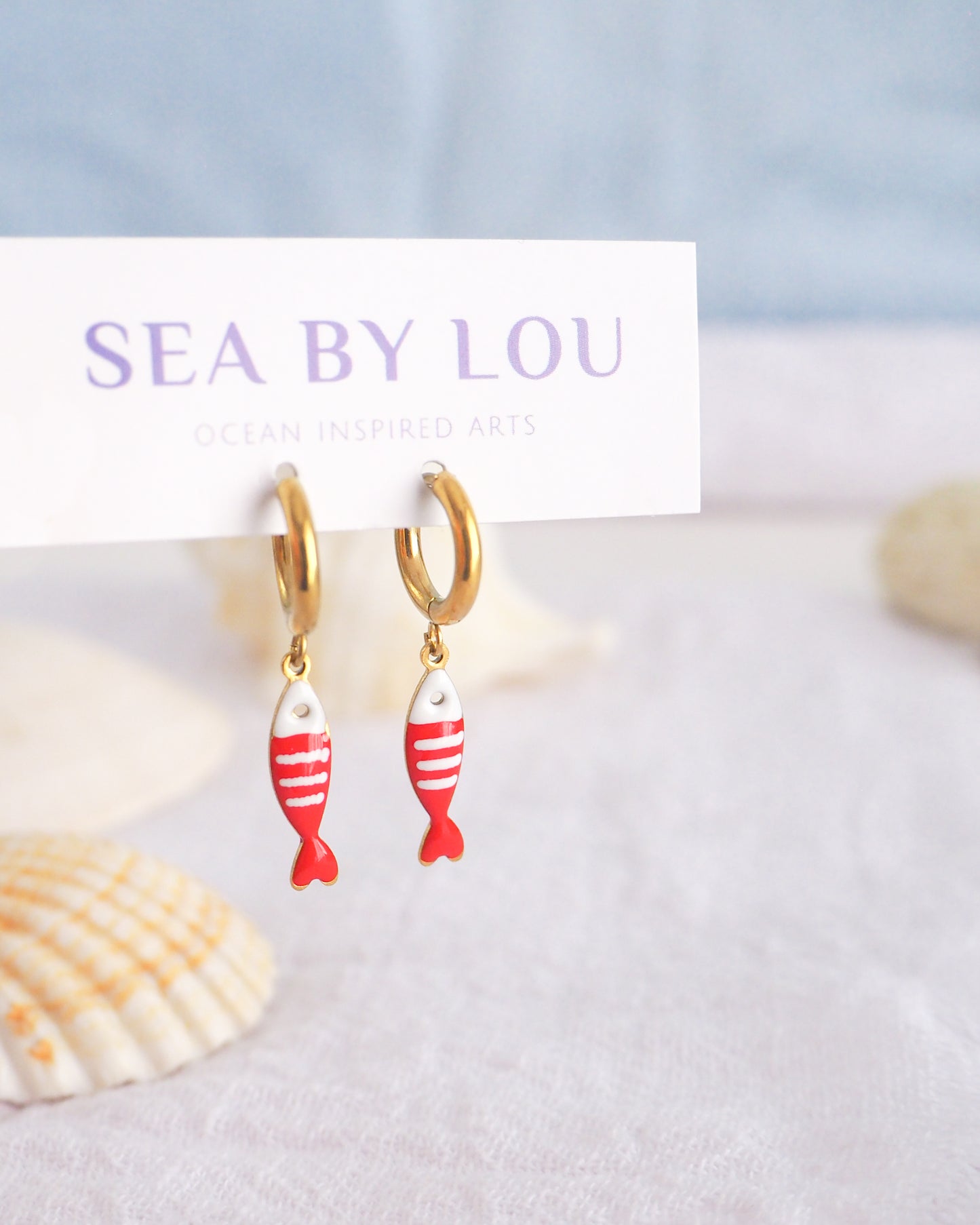 Front view of tiny sardine fish hoop earrings displayed on white Sea by Lou jewelry card. The fish pendant is red and white striped. Made with 18k gold stainless steel earring hoops. Beautiful gift for Portugal lovers or holiday memory, coastal jewelry from Portugal. Nautical minimalistic jewelry 
