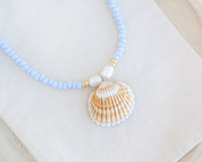Soft blue necklace with big shell pendant on white cotton displayed. The necklace is made with soft blue seed beads and freshwater pearls and a big mediterranean cockle shell. The shell is handpicked from Portugal and is a clam shaped shell with white and orange yellow lines.