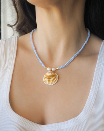 Model wearing a soft blue necklace with big shell pendant. The necklace is made with soft blue seed beads and freshwater pearls and a big mediterranean cockle shell. The shell is handpicked from Portugal and is a clam shaped shell with white and orange yellow lines. 