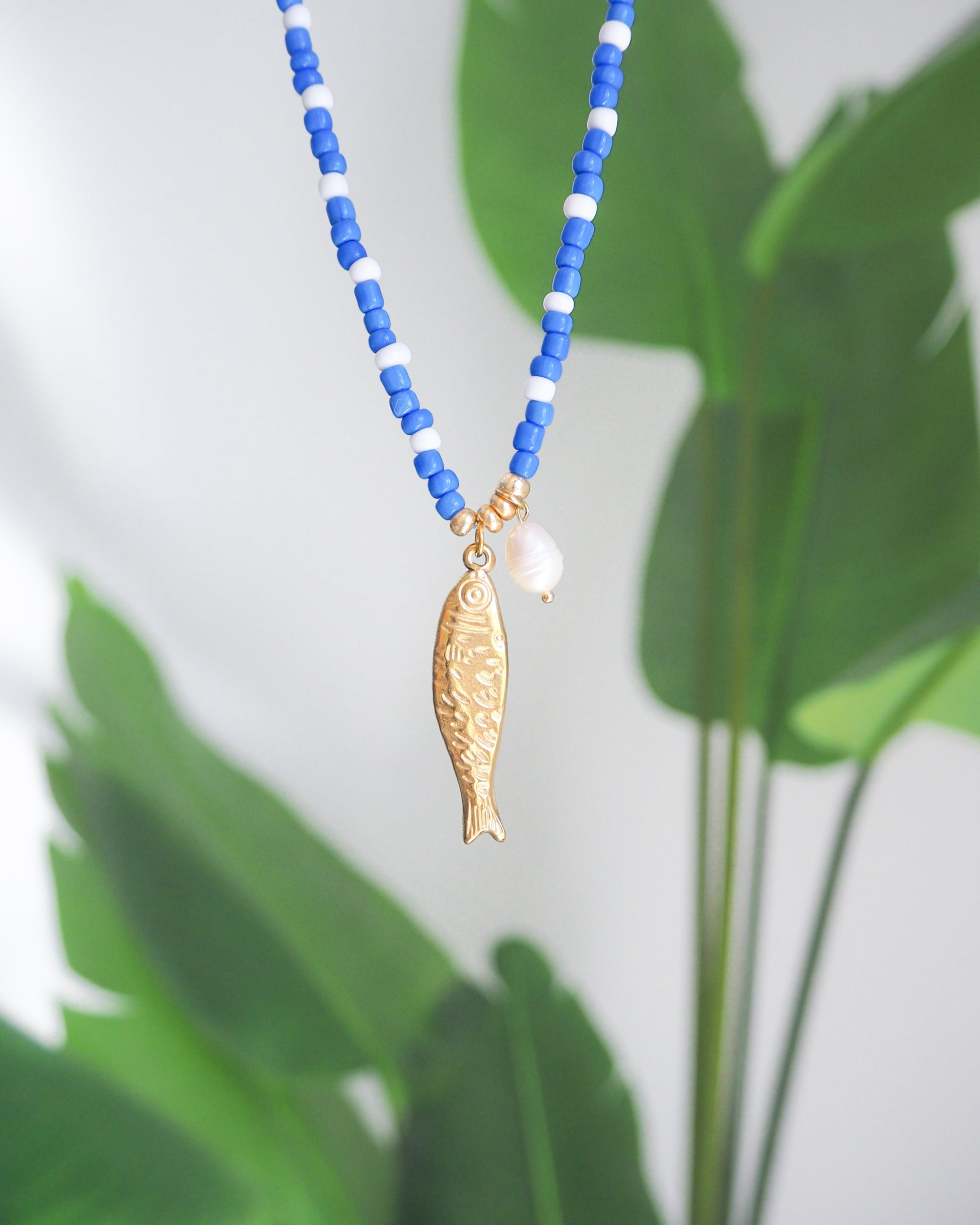 Necklace with blue and white beads and a gold fish pendant against a green leaf background. The necklace is made with seed beads and freshwater pearls and a big gold sardine fish The sardine fish pendant is made of 18k gold stainless steel. 