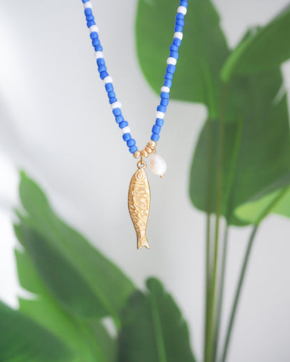 Necklace with blue and white beads and a gold fish pendant against a green leaf background. The necklace is made with seed beads and freshwater pearls and a big gold sardine fish The sardine fish pendant is made of 18k gold stainless steel. 