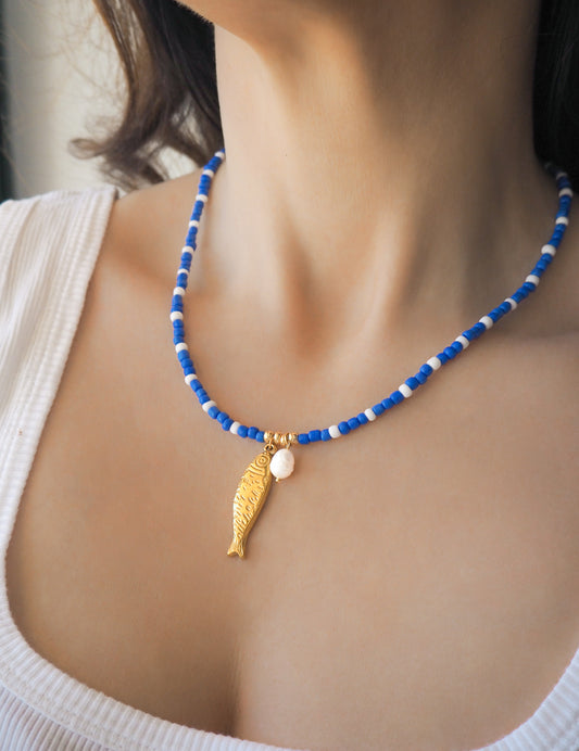 Necklace with blue and white beads and a gold sardine fish pendant and freshwater pearl bead on a person wearing a white top. The necklace is made with seed beads and freshwater pearls and a big gold sardine fish The sardine fish pendant is made of 18k gold stainless steel. 