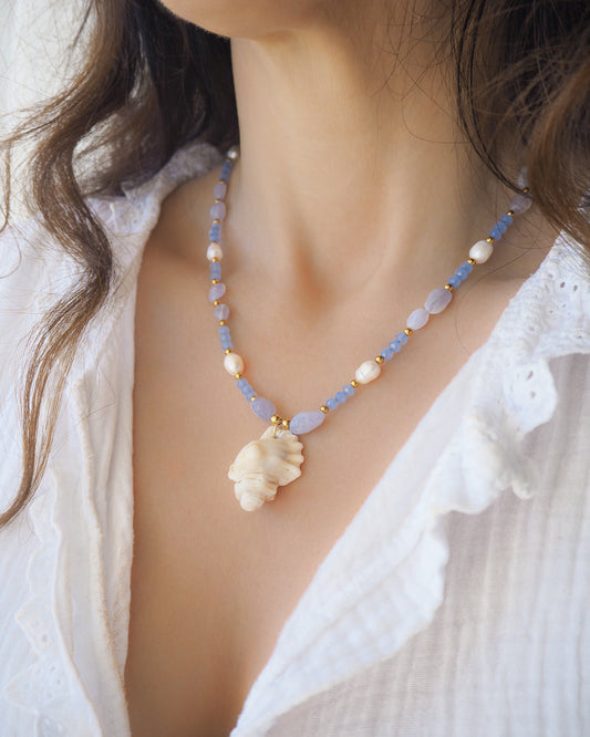 Necklace with a shell pendant worn by a person against a white background. Handmade with a real ivory mediterranean triton shell handpicked from Portugal, blue lace agate gemstone beads, blue sparkling crystal beads, ivory freshwater pearls and 18k gold stainless steel beads. The necklace is inspired on the ocean is part of the ocean serenity pearl collection. 