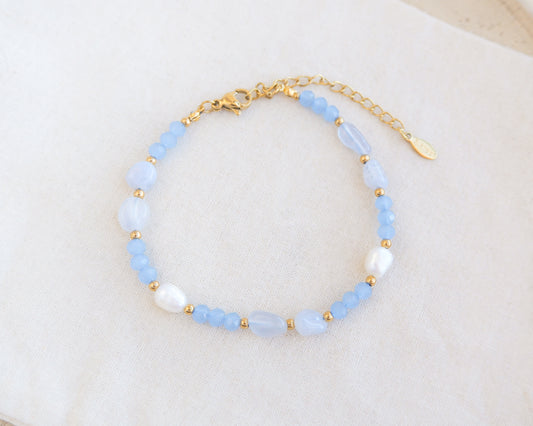 Beaded bracelet with blue and gold accents on a white background. Beaded bracelet with a white cotton background. Handmade with blue lace agate gemstone beads, blue sparkling crystal beads, ivory freshwater pearls and 18k gold stainless steel beads. The bracelet is inspired on the ocean is part of the ocean serenity pearl collection. 