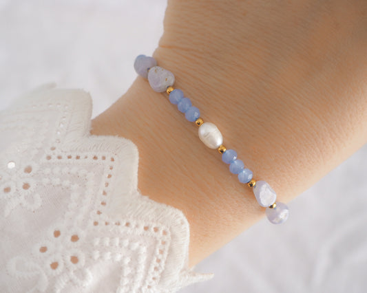 Beaded bracelet on a wrist with a white lace garment in the background. Handmade with blue lace agate gemstone beads, blue sparkling crystal beads, ivory freshwater pearls and 18k gold stainless steel beads. The bracelet is inspired on the ocean is part of the ocean serenity pearl collection. 