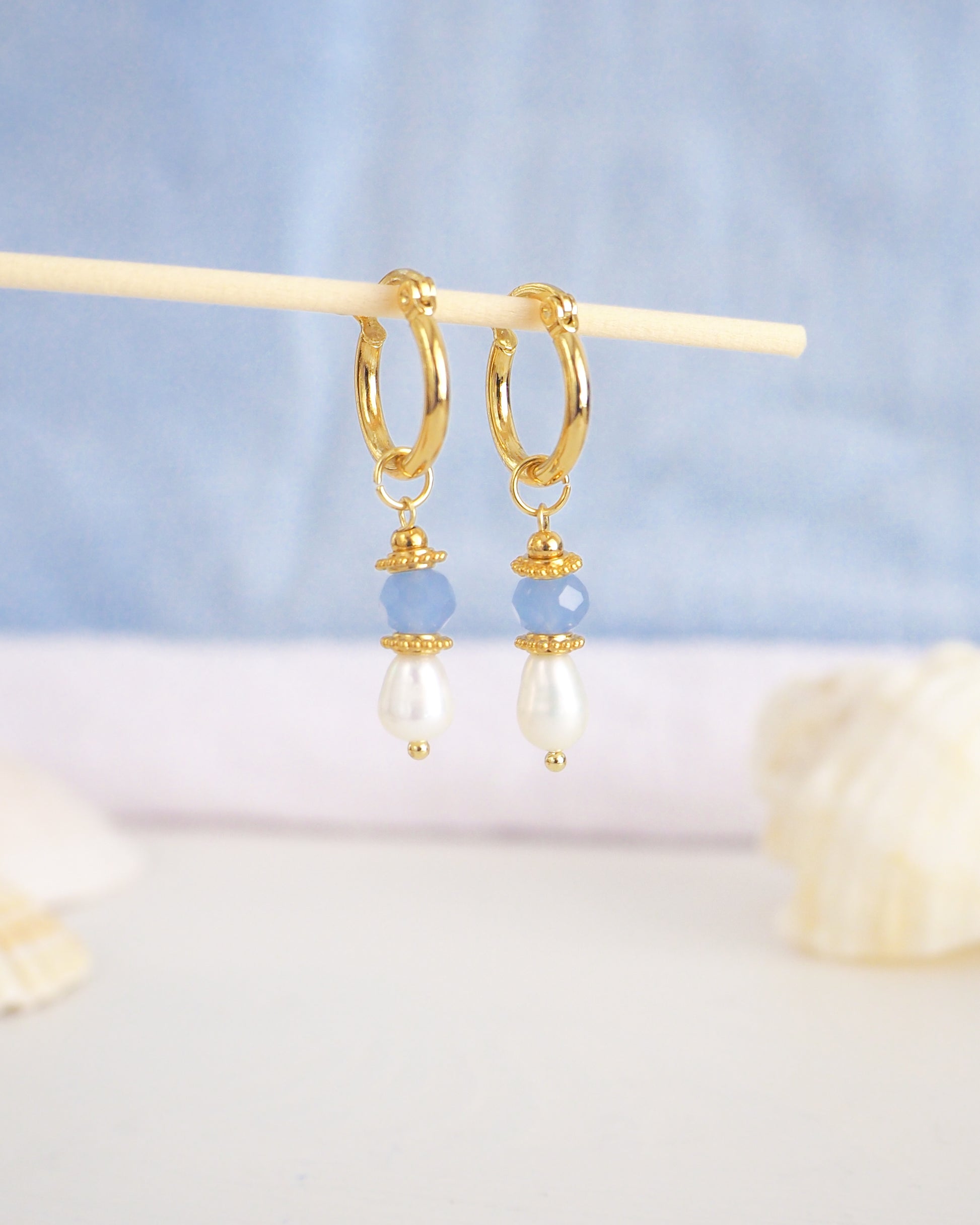 Gold hoop earrings with pearls and blue beads on a light blue background. Handmade with blue sparkling crystal beads, ivory freshwater pearls and 18k gold stainless steel beads. These earrings are inspired on the ocean is part of the ocean serenity pearl collection. 