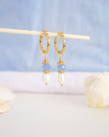 Gold hoop earrings with pearls and blue beads on a light blue background. Handmade with blue sparkling crystal beads, ivory freshwater pearls and 18k gold stainless steel beads. These earrings are inspired on the ocean is part of the ocean serenity pearl collection. 