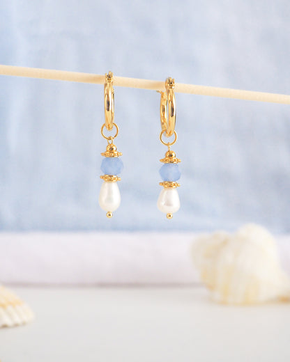 Gold hoop earrings with pearl and blue bead drops on a light background. Handmade with blue sparkling crystal beads, ivory freshwater pearls and 18k gold stainless steel beads. These earrings are inspired on the ocean is part of the ocean serenity pearl collection. 