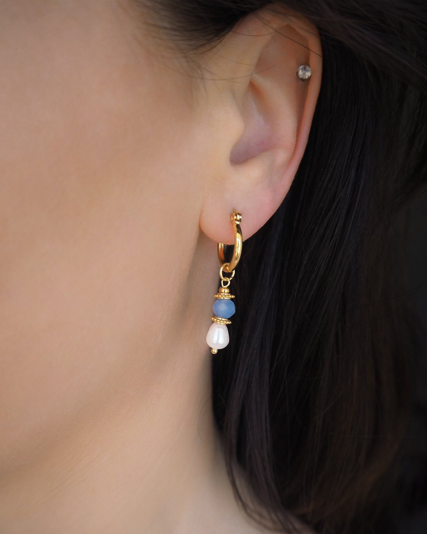 Close-up of an ear wearing a gold hoop earring with blue beads and white freshwater pearls. Handmade with blue sparkling crystal beads, ivory freshwater pearls and 18k gold stainless steel beads. These earrings are inspired on the ocean is part of the ocean serenity pearl collection. 