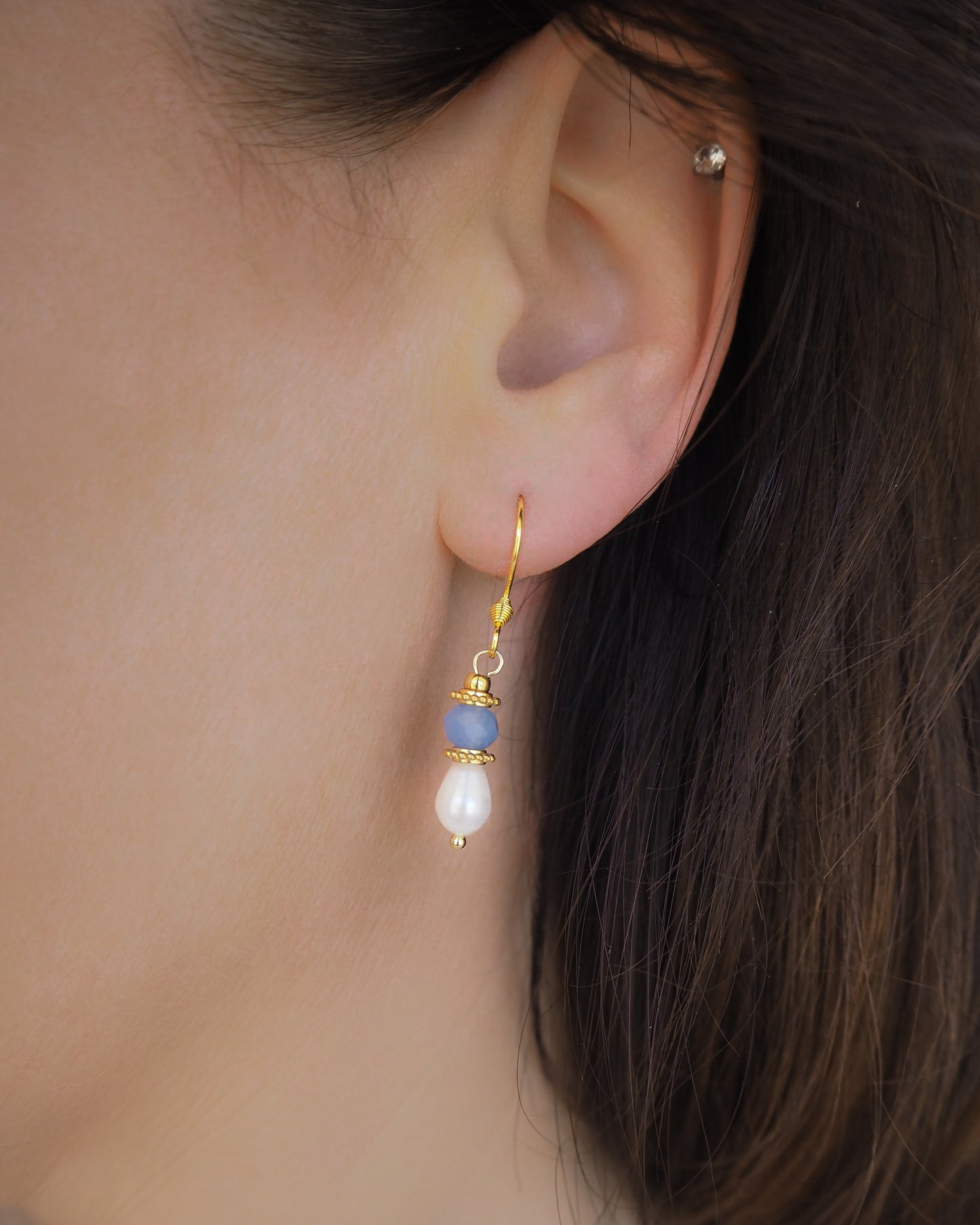 Close-up of an ear wearing a gold earring with blue and white pearls. Handmade with blue sparkling crystal beads, ivory freshwater pearls and 18k gold stainless steel beads. These earrings are inspired on the ocean is part of the ocean serenity pearl collection. 