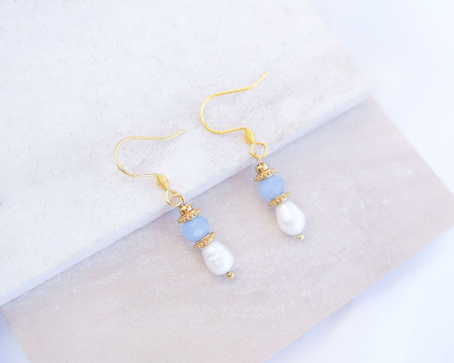 Pair of earrings with gold hooks, blue and white beads on a light background. Handmade with blue sparkling crystal beads, ivory freshwater pearls and 18k gold stainless steel beads. These earrings are inspired on the ocean is part of the ocean serenity pearl collection. 