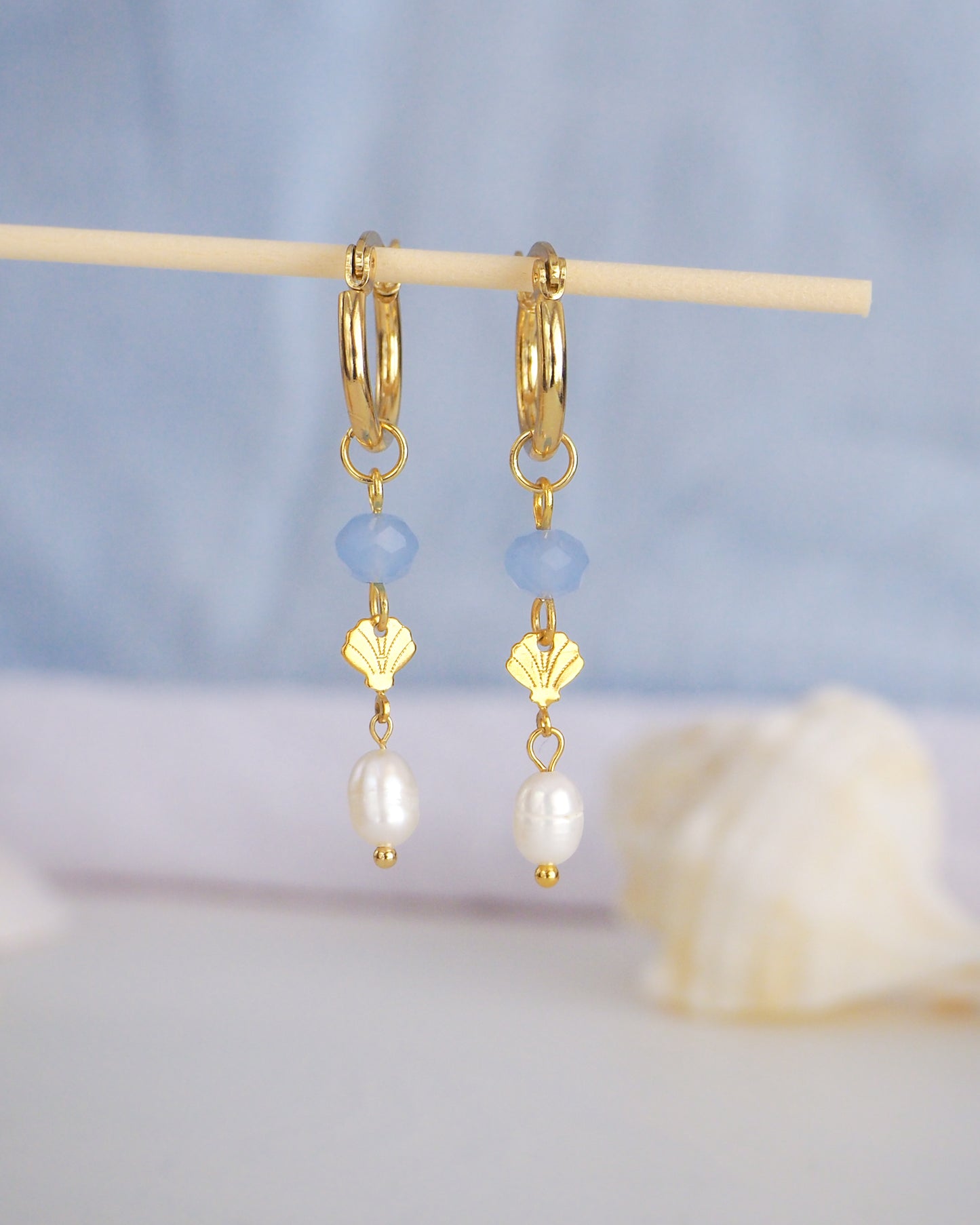 Gold hoop earrings with blue beads and pearl drops on a stick against a soft blue background. Handmade with blue sparkling crystal beads, ivory freshwater pearls and 14k gold stainless steel shell charms. The hoop earrings are made of 14k gold stainless steel These earrings are inspired on the ocean is part of the ocean serenity pearl collection.