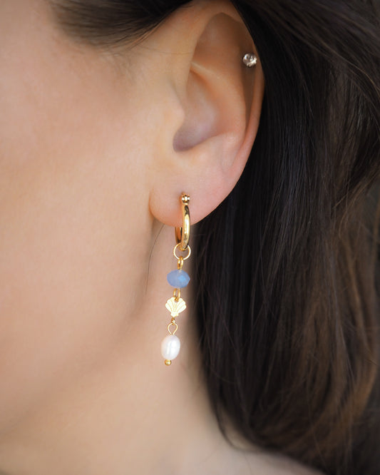 Model wearing gold hoop earrings with blue and white beads and gold shell charms in ear. Handmade with blue sparkling crystal beads, ivory freshwater pearls and 14k gold stainless steel shell charms. The hoop earrings are made of 14k gold stainless steel These earrings are inspired on the ocean is part of the ocean serenity pearl collection.