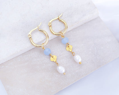 Gold hoop earrings with blue and white beads and gold shell charms on a light background. Handmade with blue sparkling crystal beads, ivory freshwater pearls and 14k gold stainless steel shell charms. The hoop earrings are made of 14k gold stainless steel These earrings are inspired on the ocean is part of the ocean serenity pearl collection.