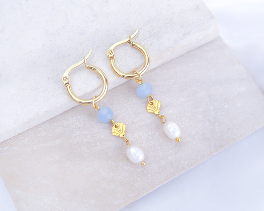Gold hoop earrings with blue and white beads and gold shell charms on a light background. Handmade with blue sparkling crystal beads, ivory freshwater pearls and 14k gold stainless steel shell charms. The hoop earrings are made of 14k gold stainless steel These earrings are inspired on the ocean is part of the ocean serenity pearl collection.