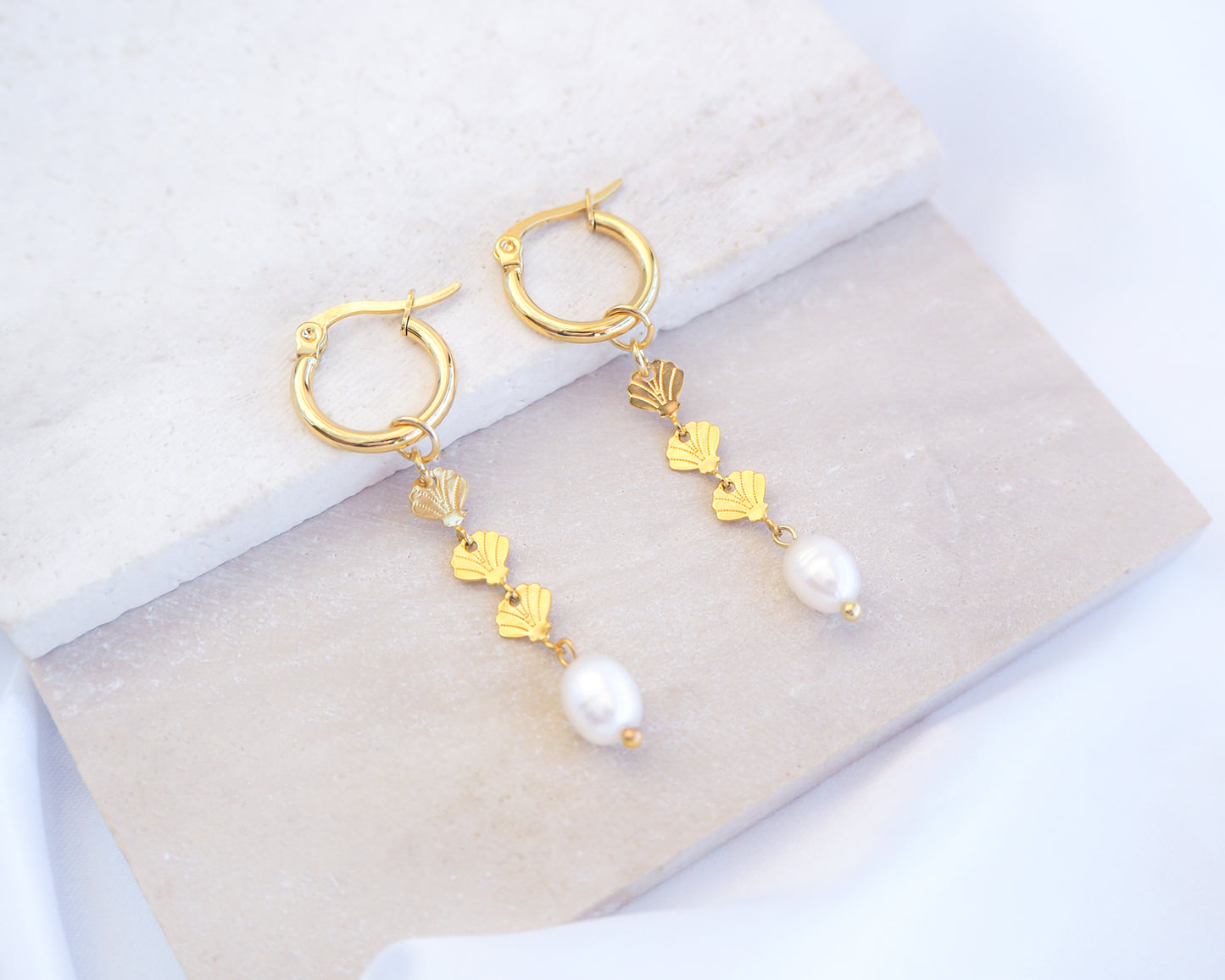 Gold hoop drop earrings with gold shells charms and freshwater pearl on a white background. The earrings are made with 14k gold stainless steel and ivory white freshwater pearls. The three shell charms are tiny and detailed. 