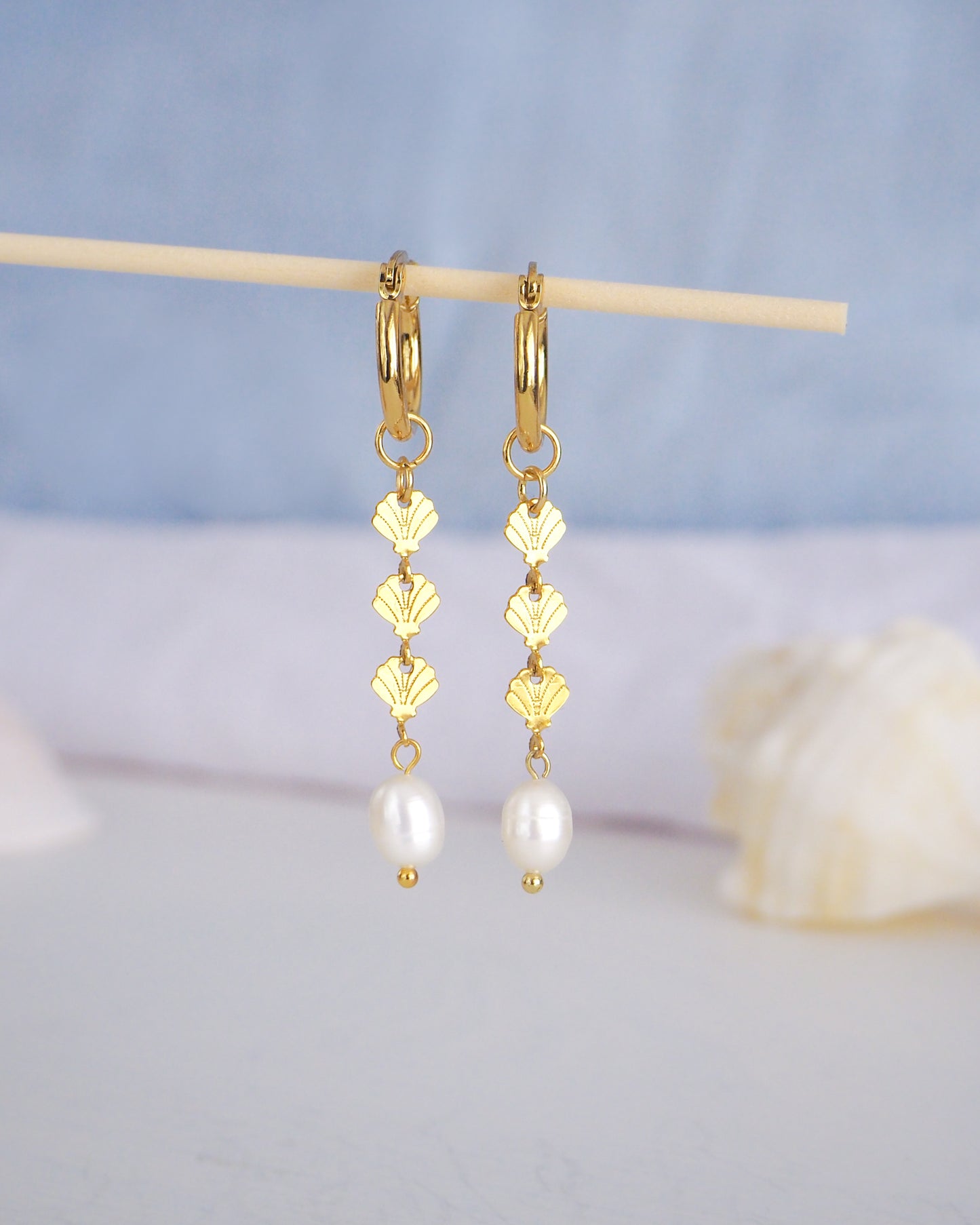 Front view of Gold hoop drop earrings with gold shells charms and freshwater pearl with blurry blue background. The earrings are made with 14k gold stainless steel and ivory white freshwater pearls. The three shell charms are tiny and detailed. 
