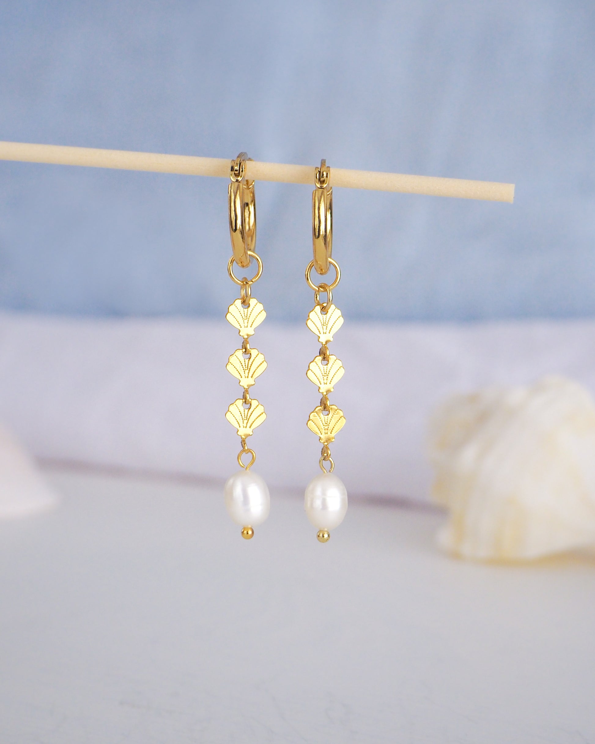Front view of Gold hoop drop earrings with gold shells charms and freshwater pearl with blurry blue background. The earrings are made with 14k gold stainless steel and ivory white freshwater pearls. The three shell charms are tiny and detailed. 