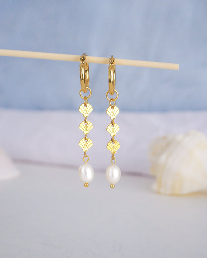 Front view of Gold hoop drop earrings with gold shells charms and freshwater pearl with blurry blue background. The earrings are made with 14k gold stainless steel and ivory white freshwater pearls. The three shell charms are tiny and detailed. 