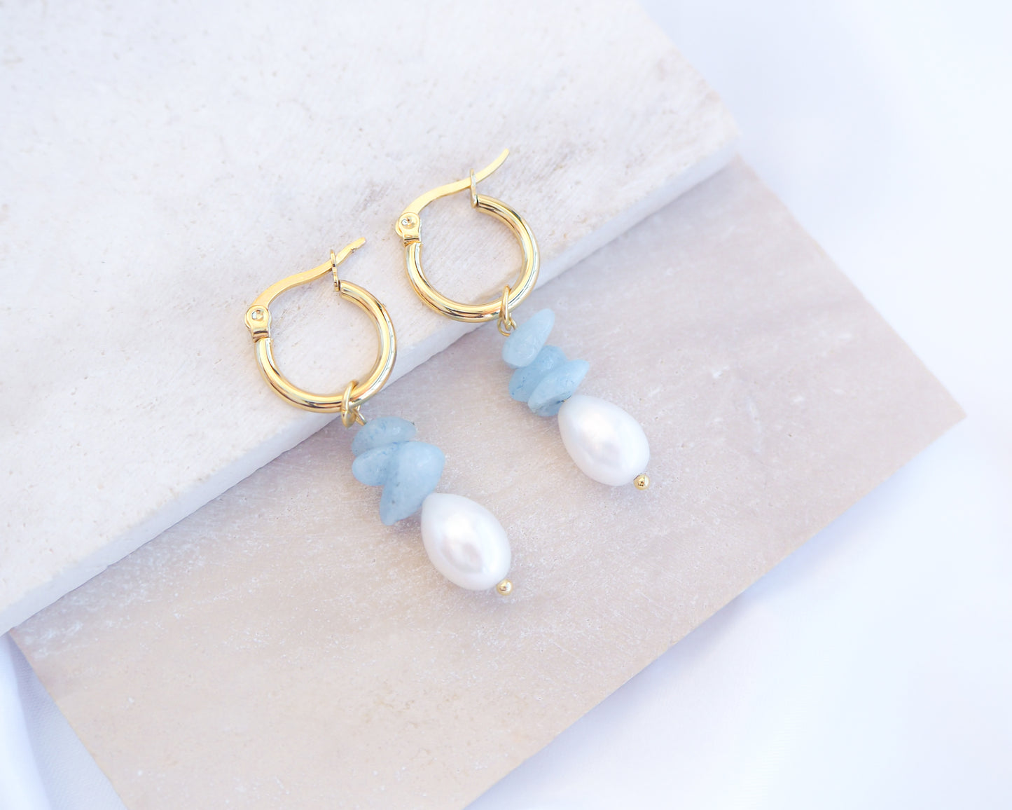 Gold hoop earrings with aquamarine gemstone beads and freshwater pearls on light grey stone displayed.