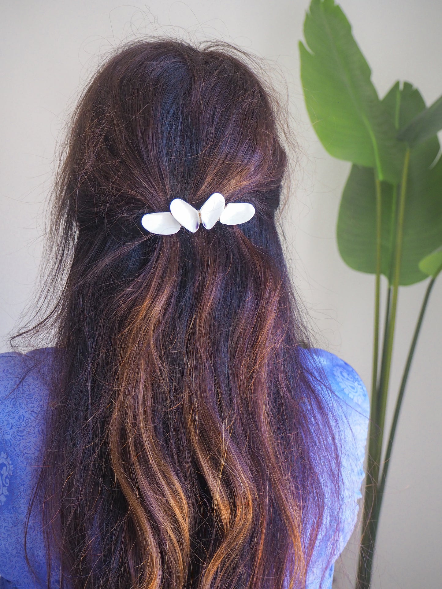 Person with wavy hair wearing a white hair clip, standing next to a plant. Model wearing White Coquina Shells Butterfly Hair Clip Barrette, Handpicked Real Coquina Shells. The shells are white in color and attached on the hair clip like a butterfly. The model has brown long hair. Sea Butterfly Shell Hair Decoration, Seashell Wedding Hair accessories, Coastal Christmas gift, Beach Wedding Hair